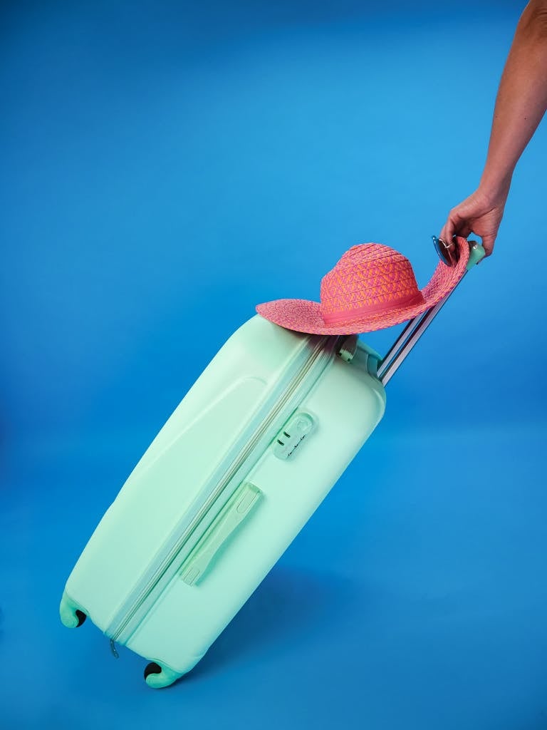 Mint green suitcase with pink sun hat against a blue background, evoking summer travel vibes.