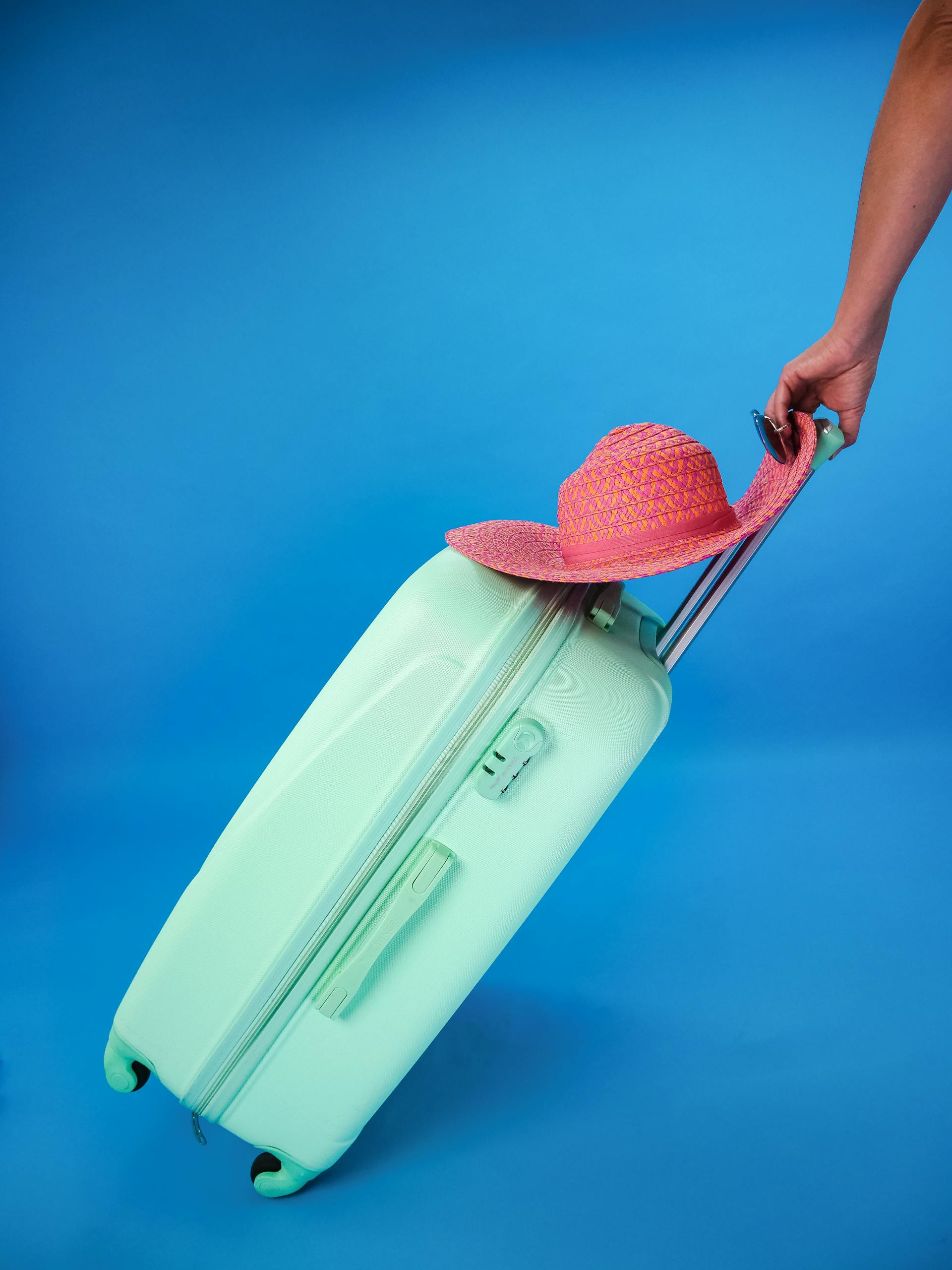 Mint green suitcase with pink sun hat against a blue background, evoking summer travel vibes.