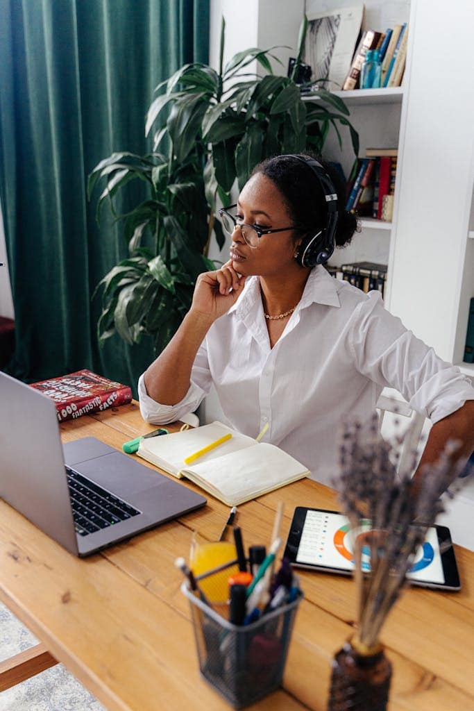 A woman in a white shirt using a laptop and tablet at a desk, wearing headphones.