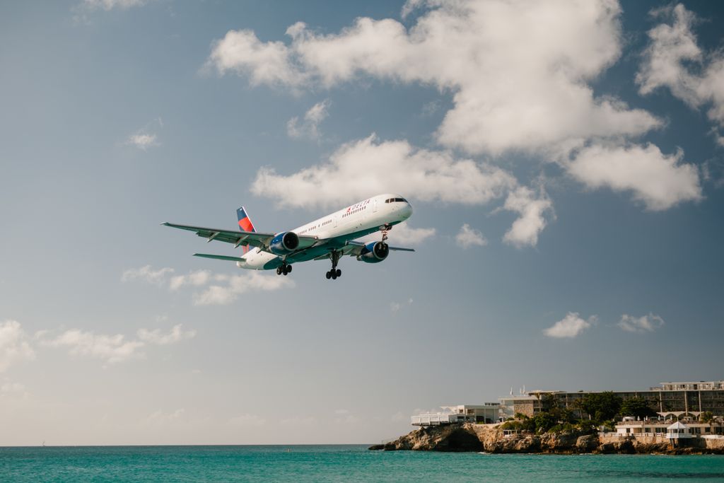 Commercial aircraft landing above ocean with clear blue skies, near beach resort.